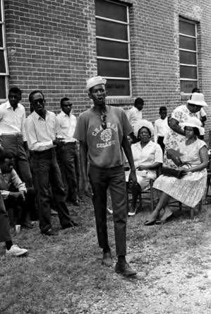 Man standing in front of an audience outside the First Missionary Baptist Church in Prattville, Alabama, during a meeting of the Autauga County Improvement Association.