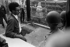 Rap Brown standing outside a fence surrounding a building in Prattville, Alabama, waiting for Stokely Carmichael's release from prison.
