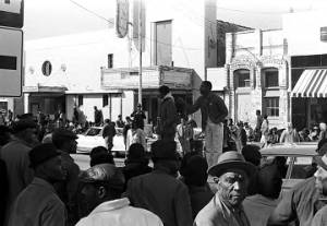 Willie Ricks standing on a car and addressing a crowd on the sidewalk in downtown Tuskegee, Alabama, during a student demonstration to protest the murder of Samuel L. Younge, a civil rights worker.