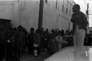Willie Ricks standing on a car and addressing a crowd on the sidewalk in downtown Tuskegee, Alabama, during a student demonstration to protest the murder of Samuel L. Younge, a civil rights worker.