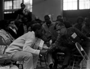 Audience listening to a young man speak while seated at the side of an auditorium or gymnasium.