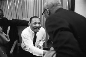 Martin Luther King, Jr., seated and shaking hands with a man during a meeting at Maggie Street Baptist Church in Montgomery, Alabama.