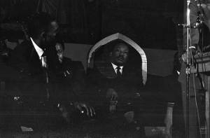 Martin Luther King, Jr., seated behind a podium and listening to Joseph Lowery speak at St. Paul AME Church in Birmingham, Alabama.