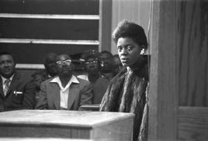 Woman in a fur jacket, seated in front of the stage of a church building, probably listening to Martin Luther King, Jr., speak.