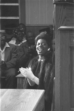 Woman in a fur jacket, seated in front of the stage of a church building, probably listening to Martin Luther King, Jr., speak.