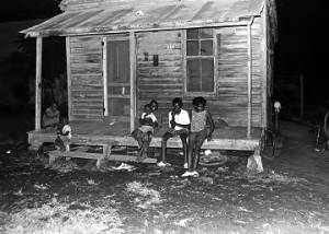 Children and young women on the front porch of a small wooden house, observing the "March Against Fear" through Mississippi at night.