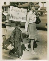 A Woman Holds A Child's Hand While Walking in Front of Demonstrators Holding a Protest Sign, circa 1970