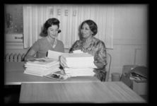Two women sitting at desk with stacks of paper and photographs in office, with the letters NEED taped on curtains in background, with box of papers on right