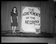 Woman with costumed child in front of sign reading "Achievements of the Negro" in front of a curtain